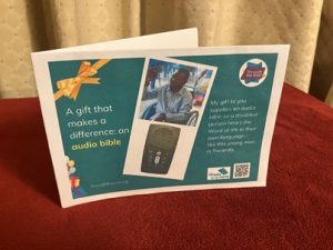 Photo of a hard copy printed gift card on a dark green background, with text on the left that says: 'A gift that makes a difference: an Audio Bible’ To the right are 2 photos: a close-up photo of a solar-powered audio Bible (which looks like a small mobile phone); a smiling young Rwandan man holding an Audio Bible up in the air, as he sits in a wheelchair. Below the images is text saying: 'My gift to you supplies an Audio Bible so a disabled person hears the Word of life in their own language - like this young man in Rwanda. There are logos for Through the Roof www.throughtheroof.org and their project Wheels for the World. There is also a QR code linking to their International Missions page at https://runyourownwebsite.uk/ttr/international-mission/