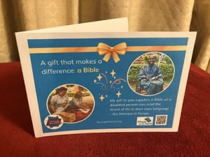 Photo of a printed gift card on a bright blue background, with text on the left that says: 'A gift that makes a difference: a Bible’ Below this is a photo of two female UK volunteers , unpacking Bibles ready to distribute. On the right is an image of a smiling young man from Kenya in a wheelchair; he wears a blue shirt and is holding a paper Bible. Below the image is text saying: 'My gift to you supplies a Bible so a disabled person can read the Word of life in their own language - like Manase in Kenya. There are logos for Through the Roof www.throughtheroof.org and their project Wheels for the World. There is also a QR code linking to their International Missions page at https://runyourownwebsite.uk/ttr/international-mission/