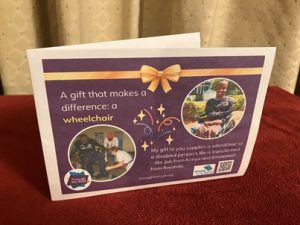 Photo of a printed gift card on a purple background, with text on the left that says: 'A gift that makes a difference: a wheelchair' Below this is a photo of a man in Rwanda being fitted with a wheelchair. On the right is an image of a smiling young man from Kenya in a wheelchair; he has no arms or legs. Below the image is text saying: 'My gift to you supplies a wheelchair so a disabled person's life is transformed - like Job in Kenya and Emmanuel in Rwanda'. showing two people receiving wheelchairs as gifts in Kenya and Rwanda. There are logos for Through the Roof www.throughtheroof.org and their project Wheels for the World. There is also a QR code linking to their International Missions page at https://runyourownwebsite.uk/ttr/international-mission/