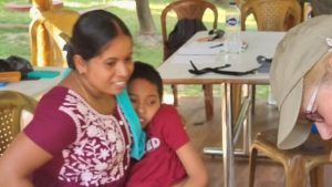 A smiling Indian mother in a purple and white sari carries her 11 year old son to a wheelchair. He is wearing a red T shirt and blue shorts.