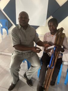 An East African man sat on a chair next to a Rwandan man seated, holding crutches, who is being presented with an audio Bible.