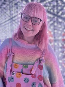 A smiling young woman with glasses and pink shoulder-length hair, wearing a dungarees with coloured spots on it.