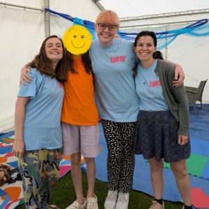 Four young women (one of whom has her face covered by a yellow smiley cartoon) all stood smiling at the camera in a marquee. The other three women are wearing light blue T shirts that say 'Team Revive' on them.