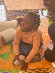 A 5 year old Kenyan girl smiling, sat on the floor with two local Kenyan volunteers.