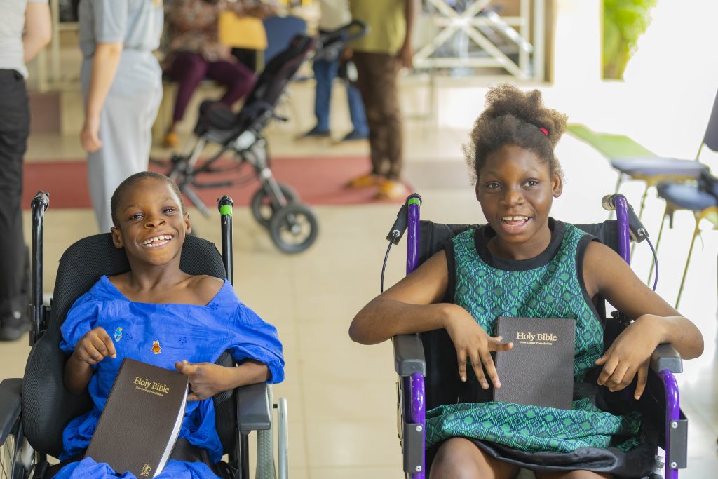 Two smiling children-in wheelchairs smiling broadly and holding Bibles in Ghana 2026