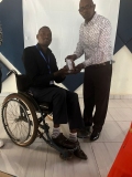 A man standing up, presenting an audio Bible to a Rwandan man in a wheelchair.