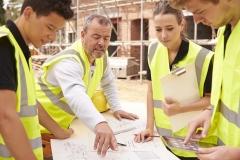 Builder On Building Site Discussing Work With Apprentice